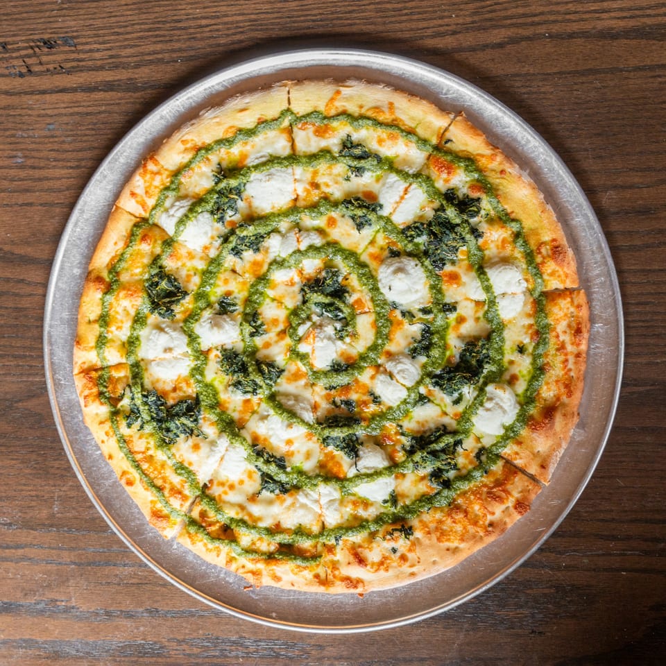 Overhead view of a sliced pizza with pesto, ricotta, and spinach on a metal tray.