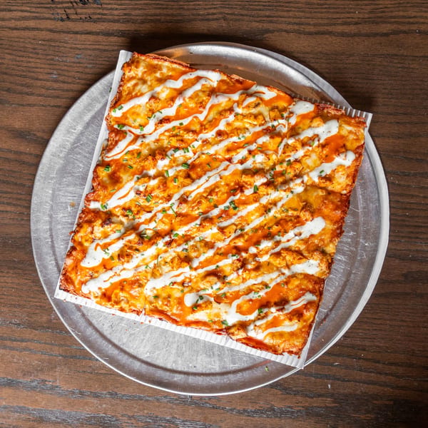 Square buffalo chicken pizza with ranch drizzle and chives on a metal tray.