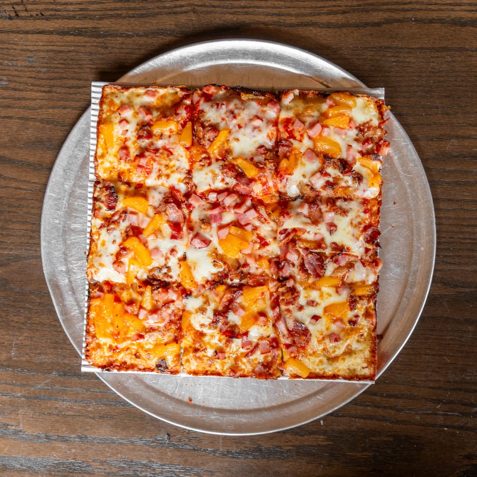 Square Detroit-style pizza with ham, bacon, and pineapple on a metal tray.