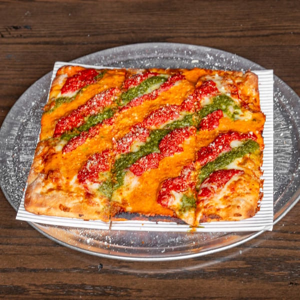 Rectangular pizza with red sauce, green pesto stripes, and grated cheese on a metal tray.