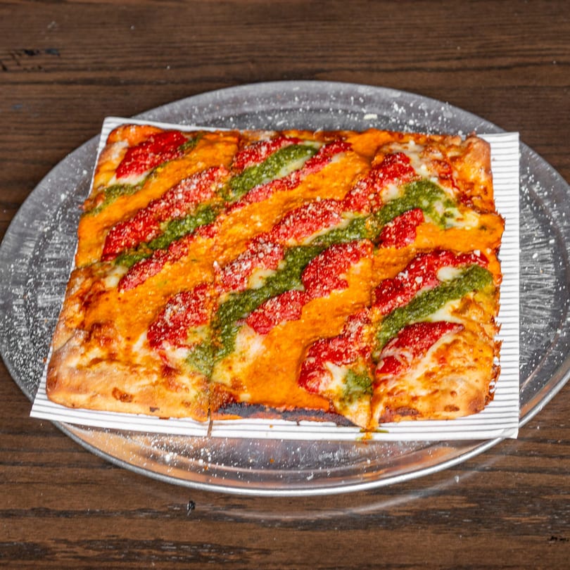 Rectangular pizza with red sauce, green pesto stripes, and grated cheese on a metal tray.